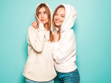 Son moda yaz kapüşonlu giysileri içinde gülümseyen iki güzel sarışın kız. Mavi duvarın yanında poz veren kaygısız seksi kadınlar. Moda ve pozitif modeller eğleniyor.