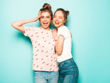 İki güzel sarışın, gülümseyen yenilikçi kız, son moda hippi kot pantolon giyiyorlar. Mavi duvarın yanında poz veren kaygısız seksi kadınlar. Moda ve pozitif modeller eğleniyor. Dillerini gösteriyorlar.