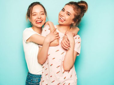 İki güzel sarışın, gülümseyen yenilikçi kız, son moda hippi kot pantolon giyiyorlar. Mavi duvarın yanında poz veren kaygısız seksi kadınlar. Moda ve pozitif modeller eğleniyor. Dillerini gösteriyorlar.