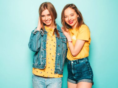 Son moda sarı tişörtlü ve kot ceketli iki güzel gülümseyen hippi kız. Mavi duvarın yanında poz veren kaygısız seksi kadınlar. Moda ve pozitif modeller eğleniyor.