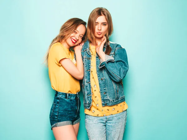 两个年轻美丽的嬉皮士女孩穿着时髦的夏季黄色T恤和牛仔裤夹克 在蓝色墙边摆姿势的无忧无虑的女人 时尚和积极的模型有乐趣 — 图库照片