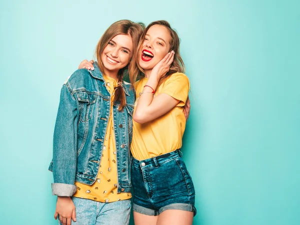 两个年轻美丽的嬉皮士女孩穿着时髦的夏季黄色T恤和牛仔裤夹克 在蓝色墙边摆姿势的无忧无虑的女人 时尚和积极的模型有乐趣 — 图库照片
