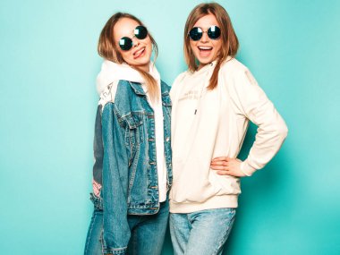 Son moda kapüşonlu ve kot ceketli gülümseyen iki güzel esmer kız. Mavi duvarın yanında poz veren kaygısız seksi kadınlar. Moda ve pozitif modeller güneş gözlükleriyle eğleniyor.