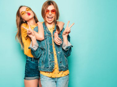 Son moda sarı tişörtlü ve kot ceketli iki güzel gülümseyen hippi kız. Mavi duvarın yanında poz veren kaygısız seksi kadınlar. Moda ve olumlu modeller barış işareti gösteriyor