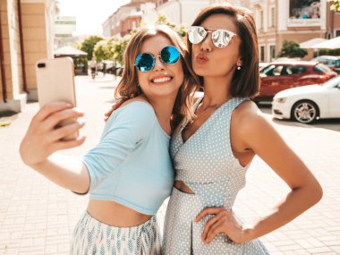 Son moda yaz kıyafetleri içinde iki güzel gülümseyen hippi kız. Sokakta güneş gözlükleriyle poz veren kaygısız kadınlar. Gün batımında akıllı telefondan kendi portresini çekiyorlar.