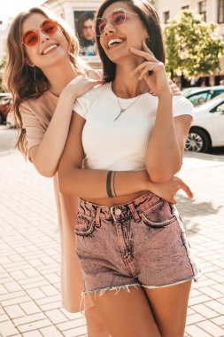 Son moda yaz kıyafetleri içinde iki güzel gülümseyen hippi kız. Güneş gözlükleriyle sokak arka planında poz veren kaygısız kadınlar. Pozitif modeller eğleniyor ve sarılıyorlar.