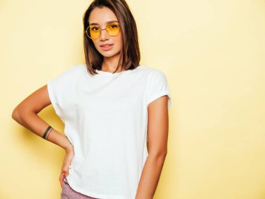 Genç ve güzel bir kadın kameraya bakıyor. Gündelik beyaz tişörtlü, kot şortlu, güneş gözlüklü, modaya uygun bir kız. Pozitif kadın yüz duyguları gösterir. Sarıda izole edilmiş komik bir model. 