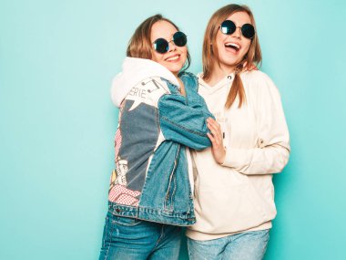 Son moda kapüşonlu ve kot ceketli gülümseyen iki güzel esmer kız. Mavi duvarın yanında poz veren kaygısız seksi kadınlar. Moda ve pozitif modeller güneş gözlükleriyle eğleniyor.