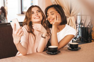 Son moda yaz kıyafetleri içinde iki güzel gülümseyen hippi kız. Kaygısız kadınlar veranda kafede sohbet ediyor ve kahve içiyor. Pozitif modeller eğleniyor ve iletişim kuruyor.