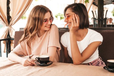Son moda yaz kıyafetleri içinde iki güzel gülümseyen hippi kız. Kaygısız kadınlar veranda kafede sohbet ediyor ve kahve içiyor. Pozitif modeller eğleniyor ve iletişim kuruyor.
