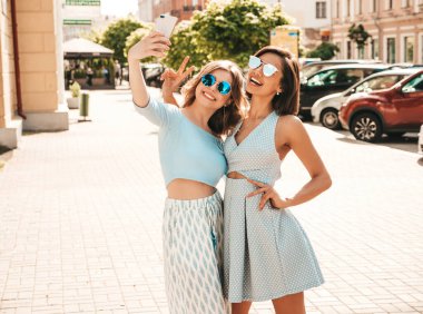 Son moda yaz kıyafetleri içinde iki güzel gülümseyen hippi kız. Sokakta güneş gözlükleriyle poz veren kaygısız kadınlar. Akıllı telefondan kendi portresini çekiyorlar. 
