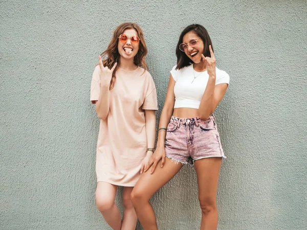 两个穿着时髦夏衣的年轻漂亮的嬉皮士女郎 戴着太阳镜 无忧无虑地躺在墙边的街上 有乐趣的正面模特 展示摇滚标志 — 图库照片