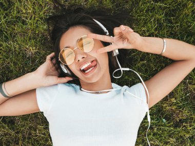 Parktaki çimlerin üzerinde yatan genç ve şık hippi kadının moda portresi. Kız modaya uygun kıyafetler giyiyor. Model barış işareti ve dil gösteriyor. Kadın kulaklıkla müzik dinliyor. Üst görünüm