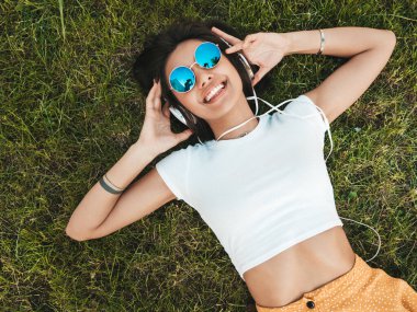 Parktaki çimlerin üzerinde yatan genç ve şık hippi kadının moda portresi. Kız modaya uygun kıyafetler giyiyor. Modeller hafta sonlarının tadını çıkarıyor. Kadın kulaklıkla müzik dinliyor. Üst görünüm