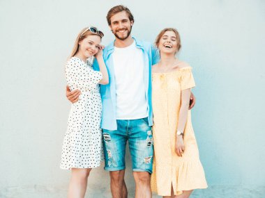 Sokakta poz veren üç genç arkadaş grubu. Moda adamı ve sıradan yaz kıyafetleri giymiş iki tatlı kız. Gülümseyen mankenler duvarın yanında eğleniyor. Neşeli kadınlar ve açık hava adamı.