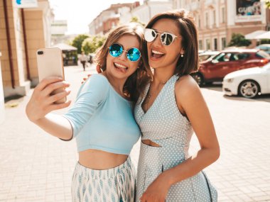 Son moda yaz kıyafetleri içinde iki güzel gülümseyen hippi kız. Sokakta güneş gözlükleriyle poz veren kaygısız kadınlar. Gün batımında akıllı telefondan kendi portresini çekiyorlar.
