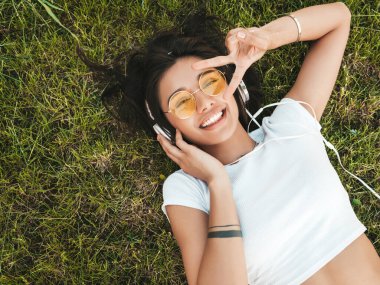 Parktaki çimlerin üzerinde yatan genç ve şık hippi kadının moda portresi. Kız modaya uygun kıyafetler giyiyor. Modeller hafta sonlarının tadını çıkarıyor. Kadın kulaklıkla müzik dinliyor. Üst görünüm