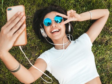 Parktaki çimlerin üzerinde yatan genç ve şık hippi kadının moda portresi. Kız modaya uygun kıyafetler giyiyor. Selfie yapıyor. Kadın kulaklıkla müzik dinliyor. Üst görünüm