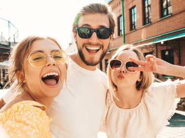Sokakta üç şık genç arkadaş grubu. Adam ve yaz kıyafetleri giymiş iki tatlı kız. Güneş gözlükleriyle eğlenen mankenler. Kadınlar ve erkek akıllı telefondan fotoğraf çektiriyorlar.