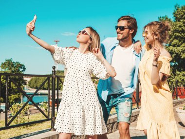 Sokakta üç şık genç arkadaş grubu. Adam ve yaz kıyafetleri giymiş iki tatlı kız. Güneş gözlükleriyle eğlenen mankenler. Kadınlar ve erkek akıllı telefondan fotoğraf çektiriyorlar.