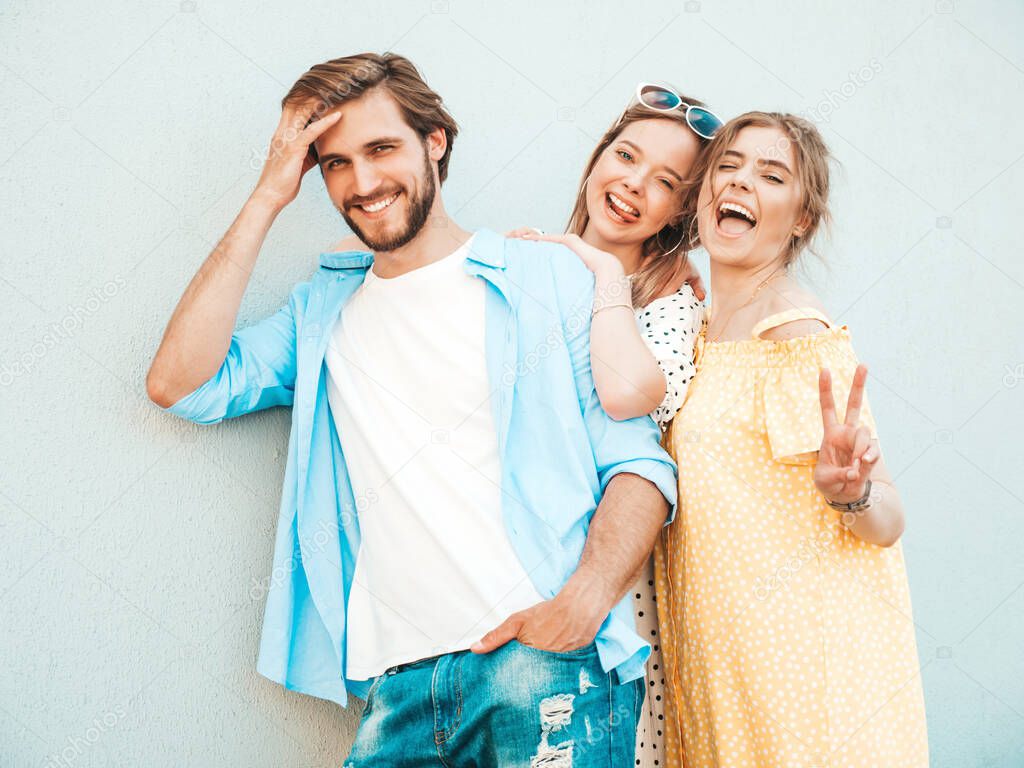 Group of young three stylish friends posing in the street. Fashion man and two cute girls dressed in casual summer clothes. Smiling models having fun near wall.Cheerful women and guy outdoors