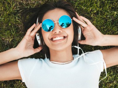 Parktaki çimlerin üzerinde yatan genç ve şık hippi kadının moda portresi. Kız modaya uygun kıyafetler giyiyor. Modeller hafta sonlarının tadını çıkarıyor. Kadın kulaklıkla müzik dinliyor. Üst görünüm