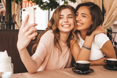 Son moda yaz kıyafetleri içinde iki güzel gülümseyen hippi kız. Kaygısız kadınlar veranda kafede sohbet edip kahve içiyor. Pozitif modeller eğleniyor ve akıllı telefondan selfie çekiyor.