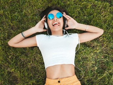Parktaki çimlerin üzerinde yatan genç ve şık hippi kadının moda portresi. Kız modaya uygun kıyafetler giyiyor. Modeller hafta sonlarının tadını çıkarıyor. Kadın kulaklıkla müzik dinliyor. Üst görünüm