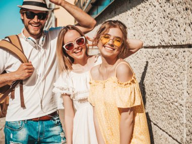 Sokakta poz veren üç genç arkadaş grubu. Moda adamı ve sıradan yaz kıyafetleri giymiş iki tatlı kız. Gülümseyen modeller güneş gözlükleriyle eğleniyorlar. Neşeli kadınlar ve açık hava adamı.