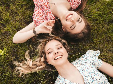 İki güzel gülümseyen yeniyetme kız şık yaz kıyafetleri içinde. Güneş gözlükleriyle yeşil çimlerde uzanan kaygısız kadınlar. Güzel mankenler.