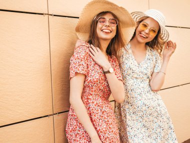 İki güzel gülümseyen yeniyetme kız şık yaz kıyafetleri içinde. Sokakta güneş gözlüklü, duvarın yanında poz veren kaygısız kadınlar. Pozitif modeller eğleniyor ve şapkalarla sarılıyorlar.