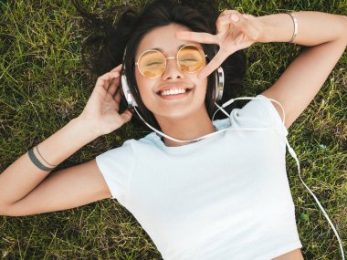 Parktaki çimlerin üzerinde yatan genç ve şık hippi kadının moda portresi. Kız modaya uygun kıyafetler giyiyor. Modeller hafta sonlarının tadını çıkarıyor. Kadın kulaklıkla müzik dinliyor. Üst görünüm
