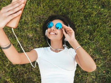 Parktaki çimlerin üzerinde yatan genç ve şık hippi kadının moda portresi. Kız modaya uygun kıyafetler giyiyor. Selfie yapıyor. Kadın kulaklıkla müzik dinliyor. Üst görünüm
