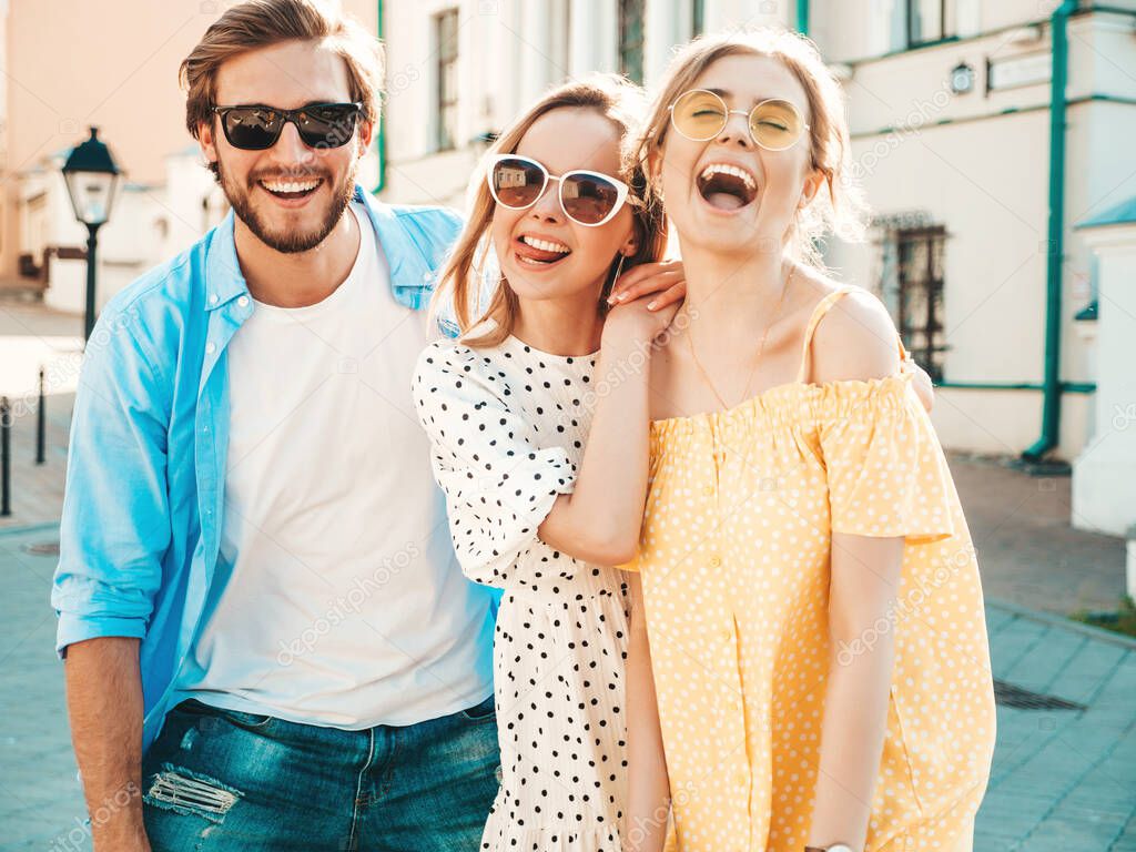 Group of young three stylish friends posing in the street. Fashion man and two cute girls dressed in casual summer clothes. Smiling models having fun in sunglasses.Cheerful women and guy at susnet
