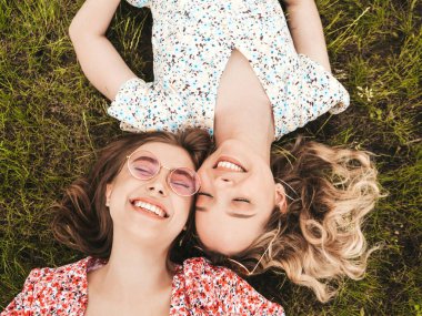 İki güzel gülümseyen yeniyetme kız şık yaz kıyafetleri içinde. Güneş gözlükleriyle yeşil çimlerde uzanan kaygısız kadınlar. Güzel mankenler.