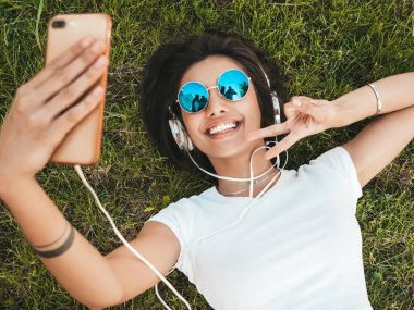 Parktaki çimlerin üzerinde yatan genç ve şık hippi kadının moda portresi. Kız modaya uygun kıyafetler giyiyor. Selfie yapıyor. Kadın kulaklıkla müzik dinliyor. Üst görünüm