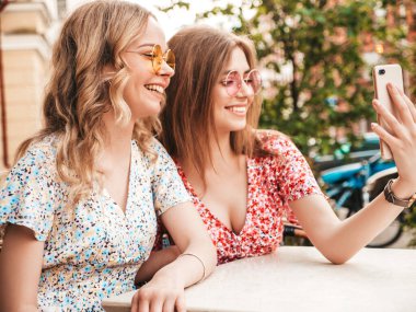 İki güzel gülümseyen hippi kız, son moda yaz kıyafetleri içinde. Kaygısız kadınlar, arka plandaki Veranda Kafe 'de sohbet ediyorlar. Pozitif mankenler eğleniyor ve akıllı telefondan selfie çekiyor.