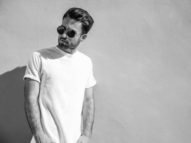 Yakışıklı, kendine güvenen, stil sahibi, Lambersex mankeni portresi. Beyaz tişört giyen adam. Moda erkeği, yuvarlak güneş gözlükleriyle duvarın yanında poz veriyor.