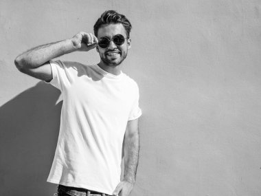 Yakışıklı, gülümseyen stil sahibi Lambersex modelinin portresi. Beyaz tişört giyen adam. Moda erkeği, yuvarlak güneş gözlükleriyle duvarın yanında poz veriyor.
