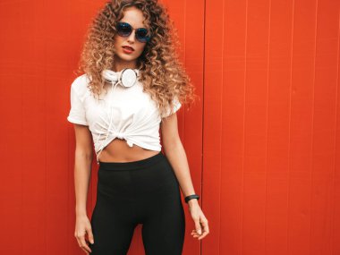 Kıvırcık saçlı güzel bir manken yaz hippi kıyafetleri içinde sokakta kırmızı duvarın yanında poz veren kaygısız bir kız. Güneş gözlükleriyle eğlenen komik ve pozitif bir kadın. 