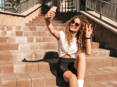 Kıvırcık saçlı güzel bir manken yaz hippi kıyafetleri giymiş, güneş gözlükleriyle sokakta poz veren kaygısız bir kız. Akıllı telefondan kendi portresini çekiyor.