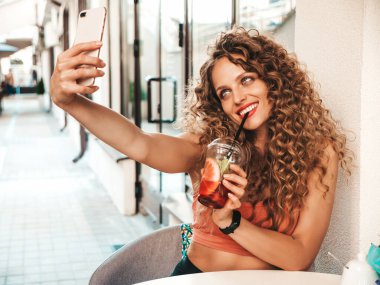 Saçları kıvırcık, genç ve güzel bir hippi kadın. Veranda kafeteryasında oturan bir kız. Modeli elinde tutarak, pipetle taze kokteyl içeceği içiyor.