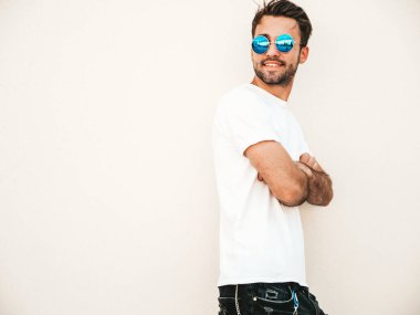 Yakışıklı, kendine güvenen, stil sahibi, Lambersex mankeni portresi. Beyaz tişört giyen adam. Moda erkeği, yuvarlak güneş gözlükleriyle duvarın yanında sokak arkasında poz veriyor.