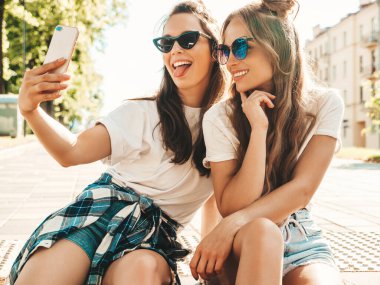 Yaz kıyafetleri içinde gülümseyen iki genç hippi kadın. Akıllı telefondan fotoğraf çeken kızlar. Sokakta oturan mankenler. Kadın çıldırıyor ve güneş gözlüklü komik suratlar yapıyor.