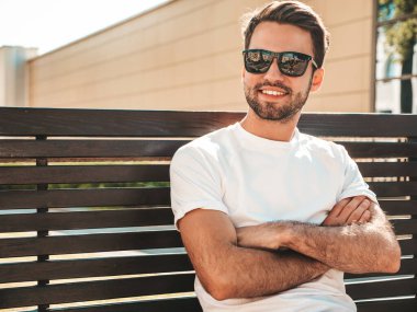 Yakışıklı, gülümseyen stil sahibi Lambersex modelinin portresi. Beyaz tişört giyen adam. Moda erkeği sokağın ortasında güneş gözlükleriyle oturuyor. Kollarını kavuşturmuş.
