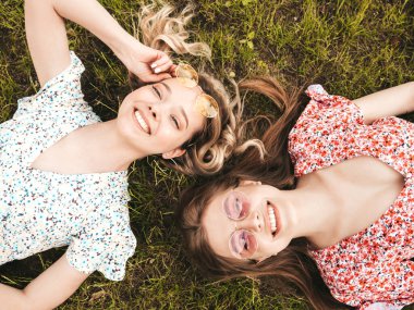 İki güzel gülümseyen yeniyetme kız şık yaz kıyafetleri içinde. Güneş gözlükleriyle yeşil çimlerde uzanan kaygısız kadınlar. Güzel mankenler.