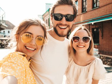 Sokakta üç şık genç arkadaş grubu. Adam ve yaz kıyafetleri giymiş iki tatlı kız. Güneş gözlükleriyle eğlenen mankenler. Kadınlar ve erkek akıllı telefondan fotoğraf çektiriyorlar.
