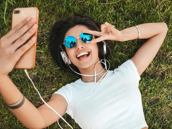 Parktaki çimlerin üzerinde yatan genç ve şık hippi kadının moda portresi. Kız modaya uygun kıyafetler giyiyor. Selfie yapıyor. Kadın kulaklıkla müzik dinliyor. Üst görünüm