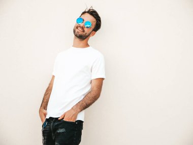 Yakışıklı, gülümseyen stil sahibi Lambersex modelinin portresi. Beyaz tişört giyen adam. Moda erkeği, yuvarlak güneş gözlükleriyle duvarın yanında sokak arkasında poz veriyor.