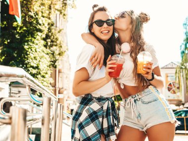 Moda kıyafetleri içinde gülümseyen iki güzel hippi kız. Dışarıda poz veren kaygısız kadınlar. Pozitif modeller, plastik bardakta pipetle taze kokteyl içiyorlar ve içiyorlar.